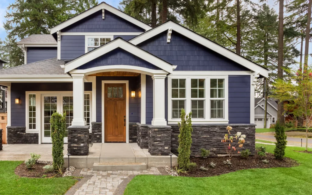 Photo of the front of a house with dark blue siding, white trim, and a large front porch with slate stone foundation.