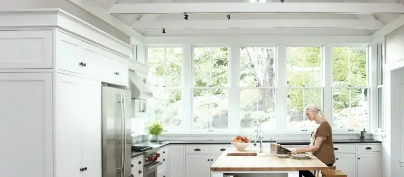 Photo of a woman sitting in her modern white kitchen going over paperwork in front of Pella windows.