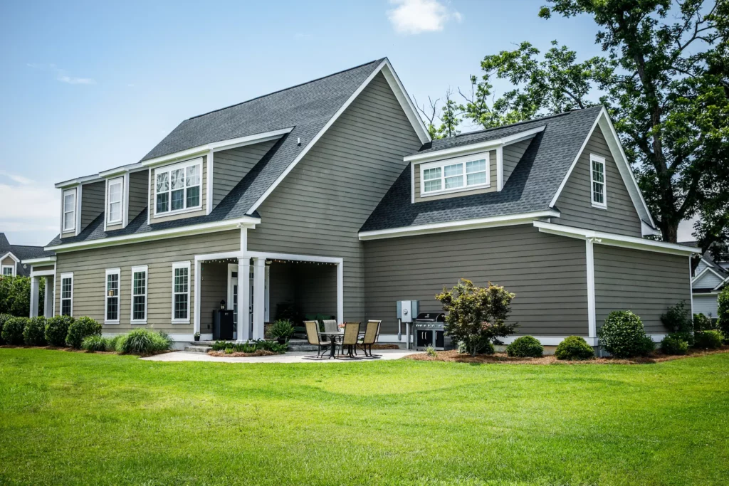 Photo of a home with gray siding, two stories, attached garage, and a partially covered back patio with table and grill. Landscaped shrubs wrap around the home.