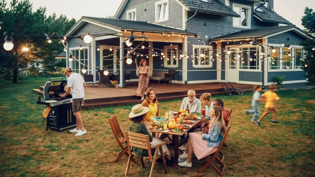 Photo of a family enjoying a cookout around a wooden table in the yard behind their home with dark blue siding and raised wooden porch. Kids are running around the yard. A light net is strung from the house out.