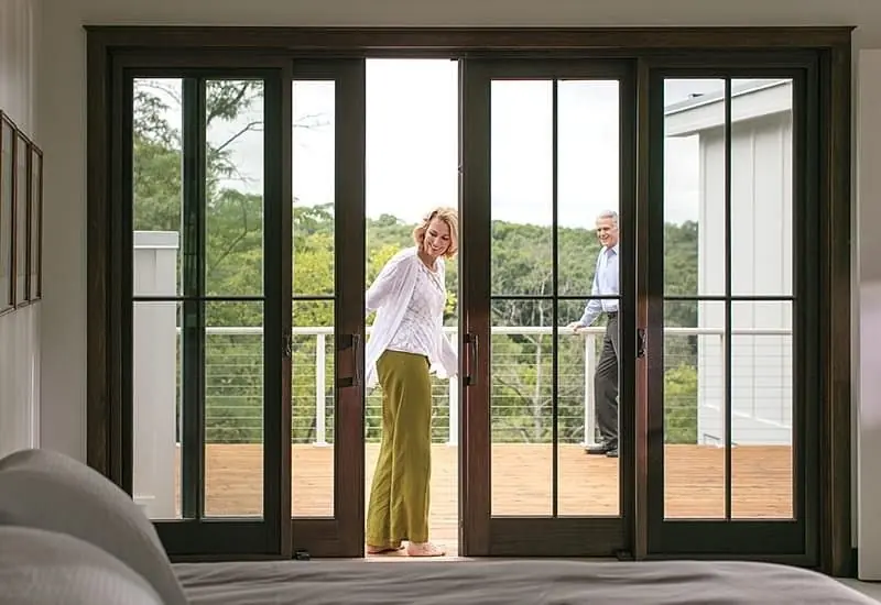Photo of people on their deck overlooking a forested area and closing a dark wood sliding glass door. Pella integrates perfectly with outdoor living.