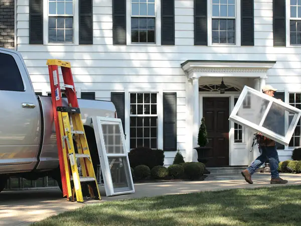 Silver pickup truck with ladders and new windows perched on it in front of a large white house with black trim. A team member is carrying one of the windows up to the house.