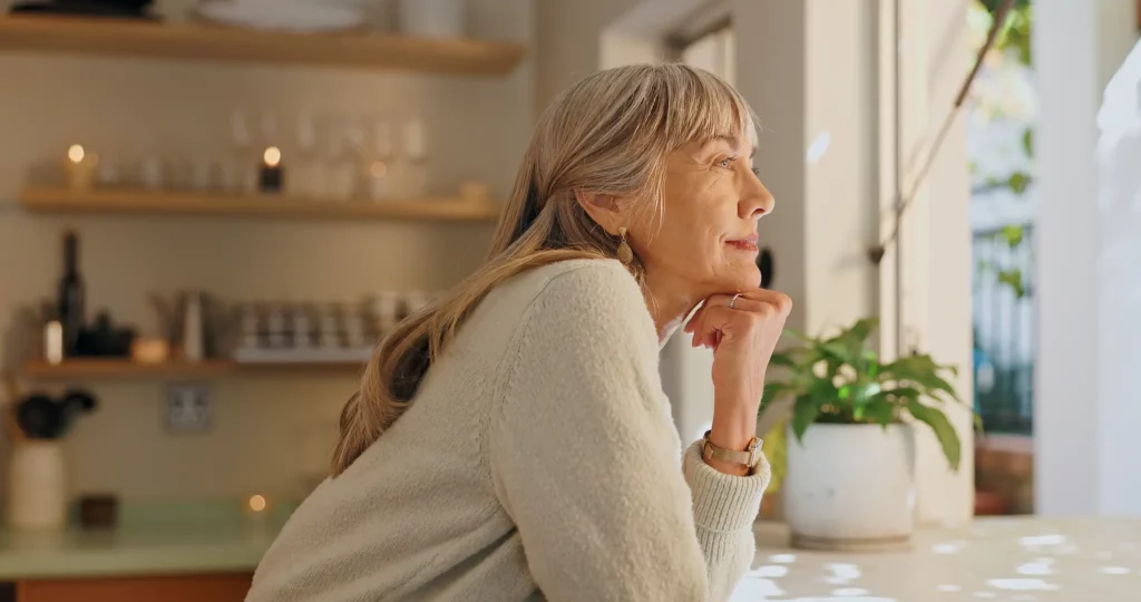 Woman gazing out her home window with her hand under her chin.