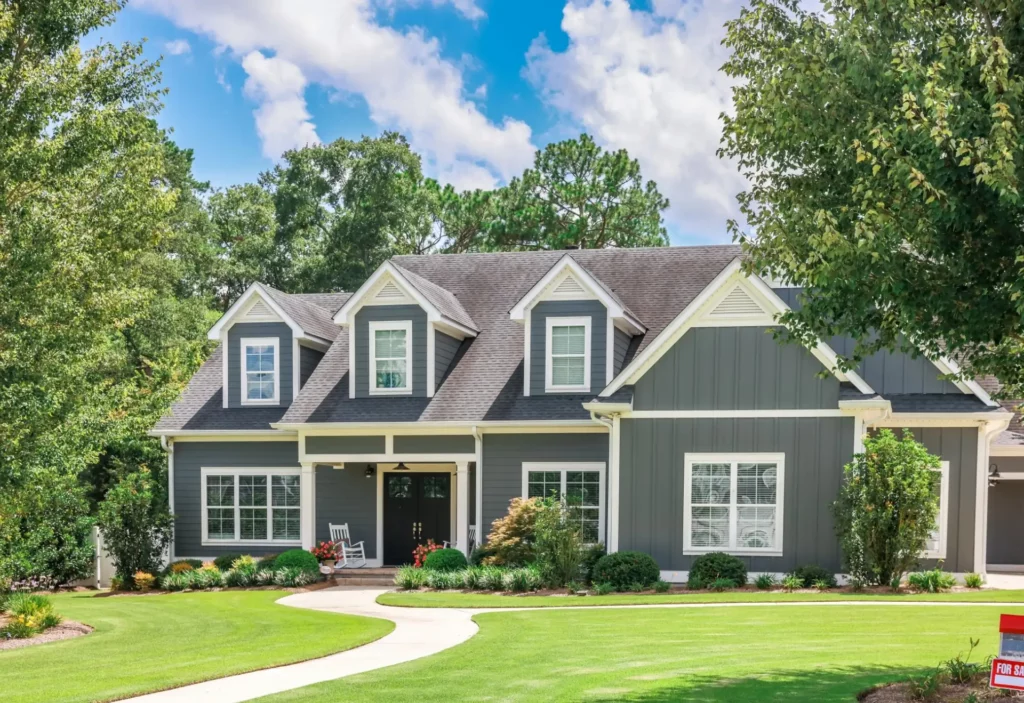 Photo of a two-story house with bluish-gray siding, white trim, and landscaping with a mix of shrubs and bushes. The path winds around from the front door to the street and over to the driveway.