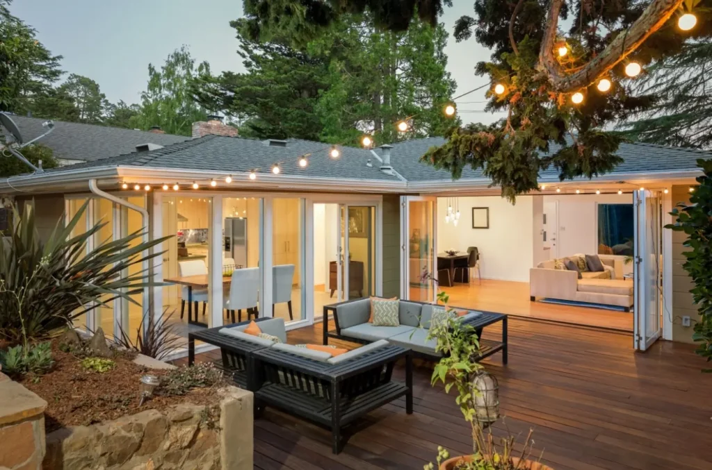 Single-story home with green siding and large patio with full-length sliding glass doors and windows. String lights are hung from the under-hang to a tree. Rock mulched flower bed.